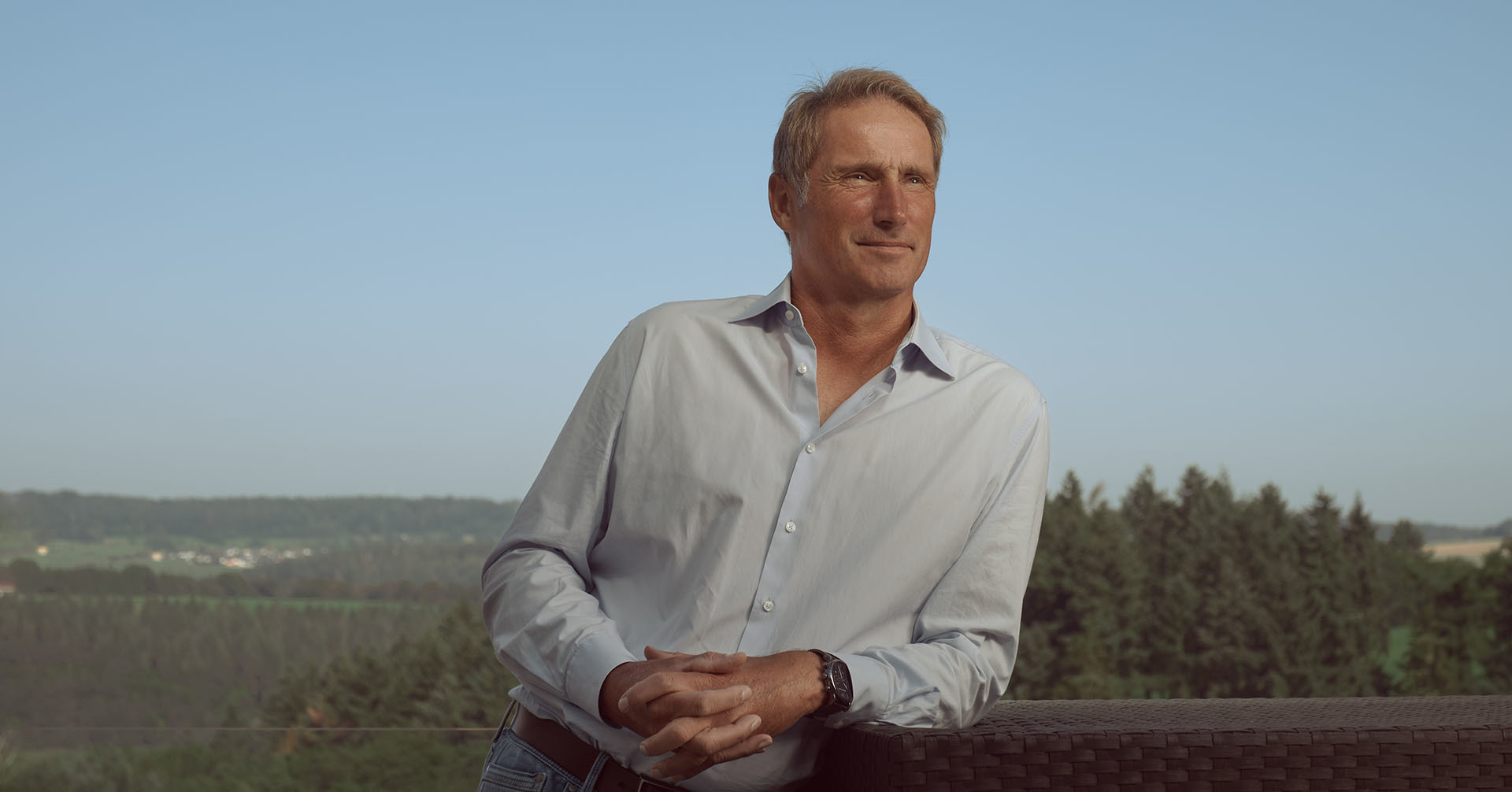 Stefan Burchard steht auf einem Tennisplatz hinter dem Netz. Er trägt eine Trainingsjacke mit den Farben Schwarz-Rot-Gold und dem Bundesadler-Emblem, hält einen Tennisschläger in der Hand und blickt ernst nach vorn. Der Himmel im Hintergrund ist leicht bewölkt.