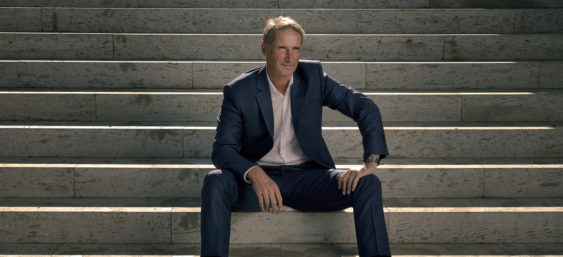 Ein Mann, Stefan Burchard, im blauen Anzug sitzt auf einer breiten Steintreppe. Er trägt ein weißes Hemd, braune Lederschuhe und eine Armbanduhr. Mit ernstem Blick schaut er in die Kamera, die Hände lässig auf den Oberschenkeln abgestützt. Die Szene ist klar und symmetrisch komponiert, das Licht fällt in Streifen über die Stufen.